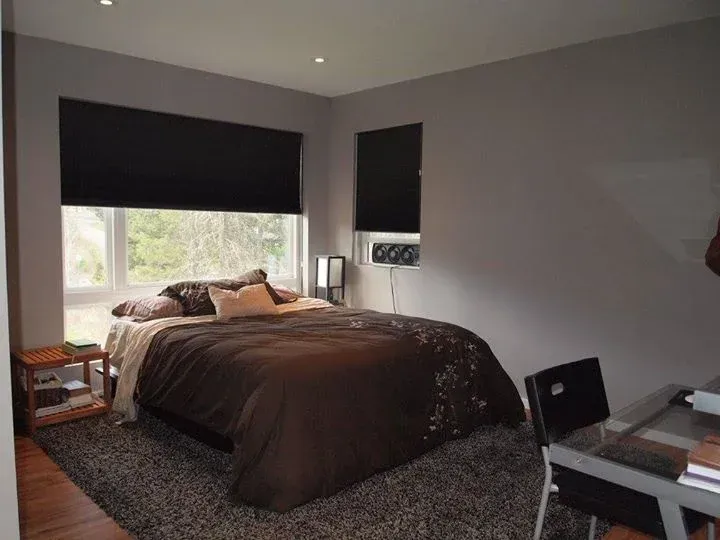 Bedroom with a bed, desk, and window with black shades; gray walls, brown bedding.