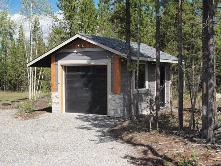Small gray garage with a dark door, stone accents, and gravel driveway.