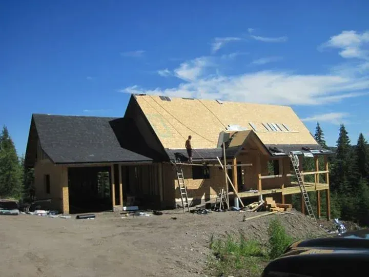 House under construction; workers on roof installing shingles. Blue sky, garage, and wooded background.