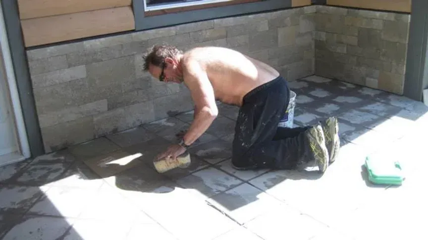 Man on knees, shirtless, laying stone patio tiles outdoors. Sunny.