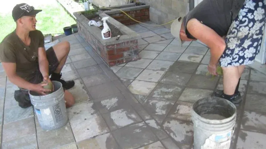 Two people apply cement grout to patio tile; one mixing in a bucket, the other cleaning the tiles.