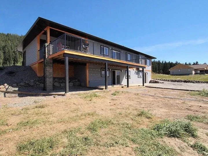 Two-story gray house with black railing, deck, and windows, on a gravel lot, under blue sky.