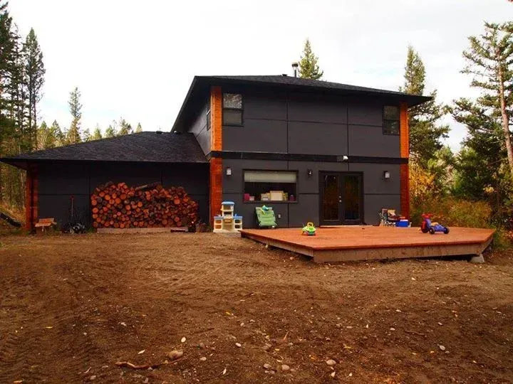 Modern dark grey house with firewood, deck, and forest backdrop.