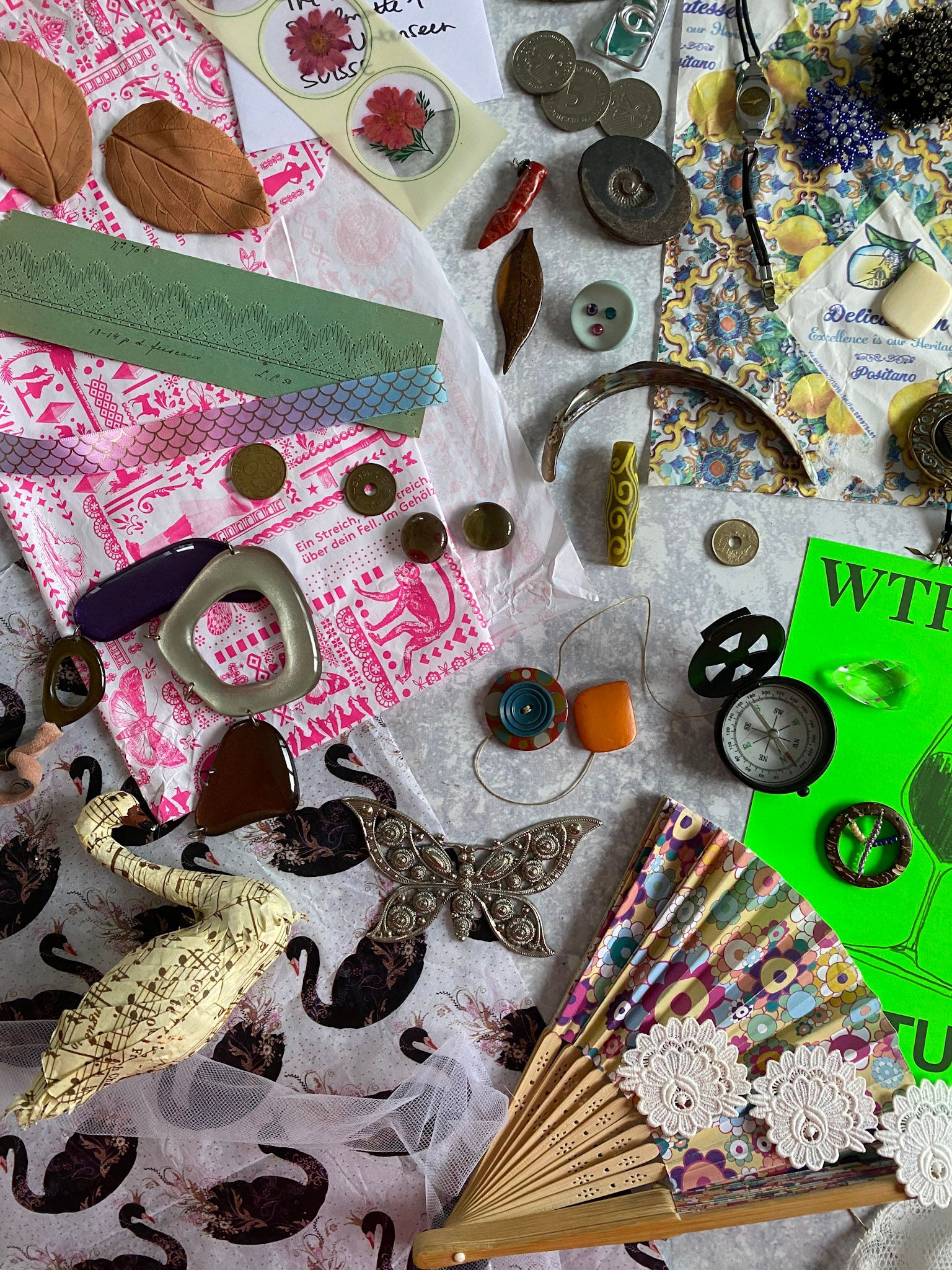 Assorted craft supplies: buttons, paper cutouts, beads, fabric scraps, and a fan, arranged on white and patterned surfaces.