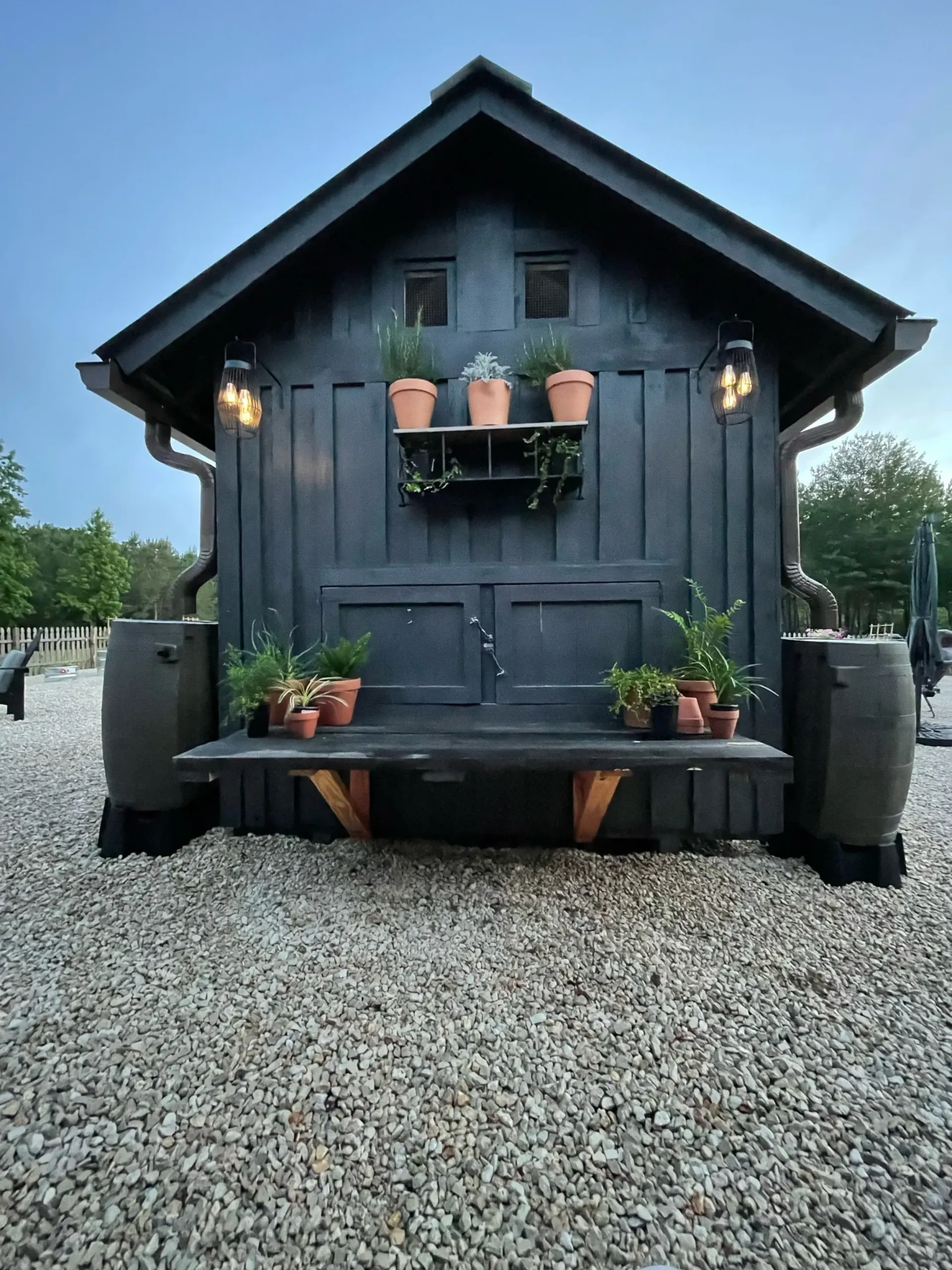 Dark gray garden shed with potted plants and bench on gravel.