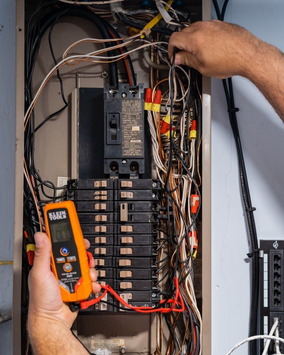 Hands testing electrical wiring inside a residential circuit breaker panel using a digital multimeter.