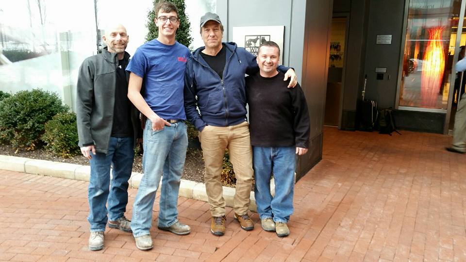 Four people stand together in a group portrait outdoors on a brick patio in front of a building entrance.