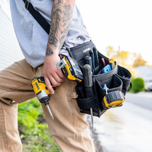 A worker in khaki pants holding a yellow power drill with a tool pouch worn at the hip, outdoors.