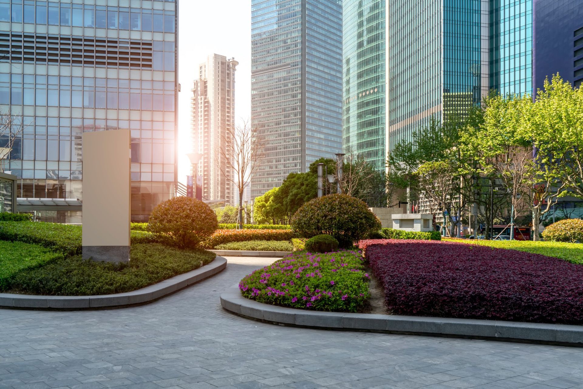 Modern skyscrapers tower over a landscaped urban plaza with paved walkways, manicured hedges, and lush green bushes.