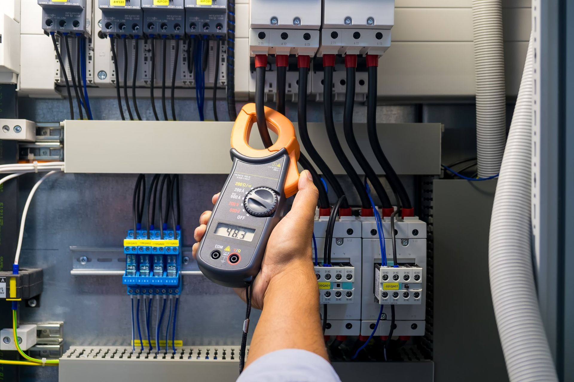 A hand uses a digital clamp meter to measure the current of a black wire inside an electrical control panel.