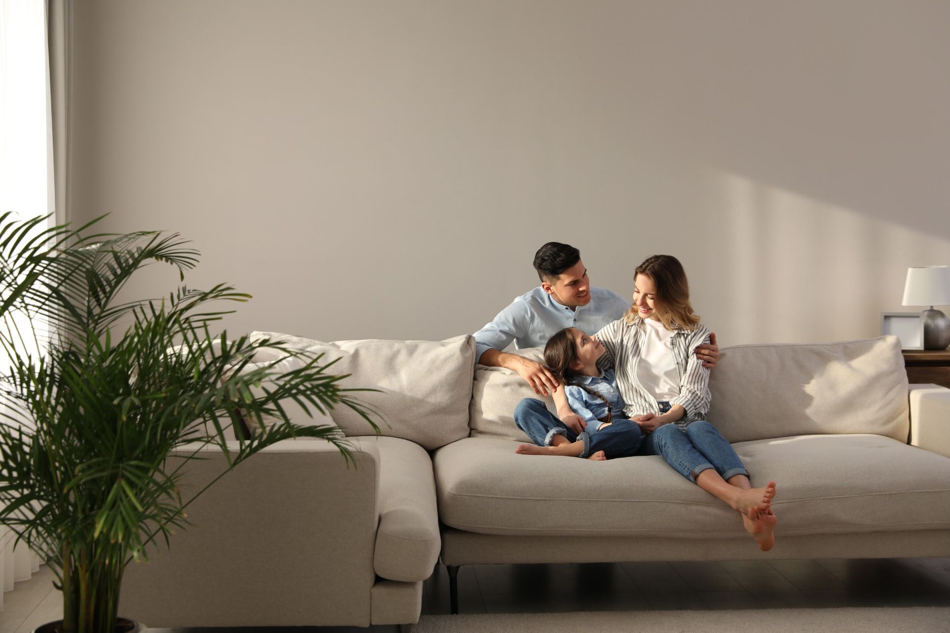 A family sitting together on a light-colored couch in a sunlit living room with a houseplant in the foreground.