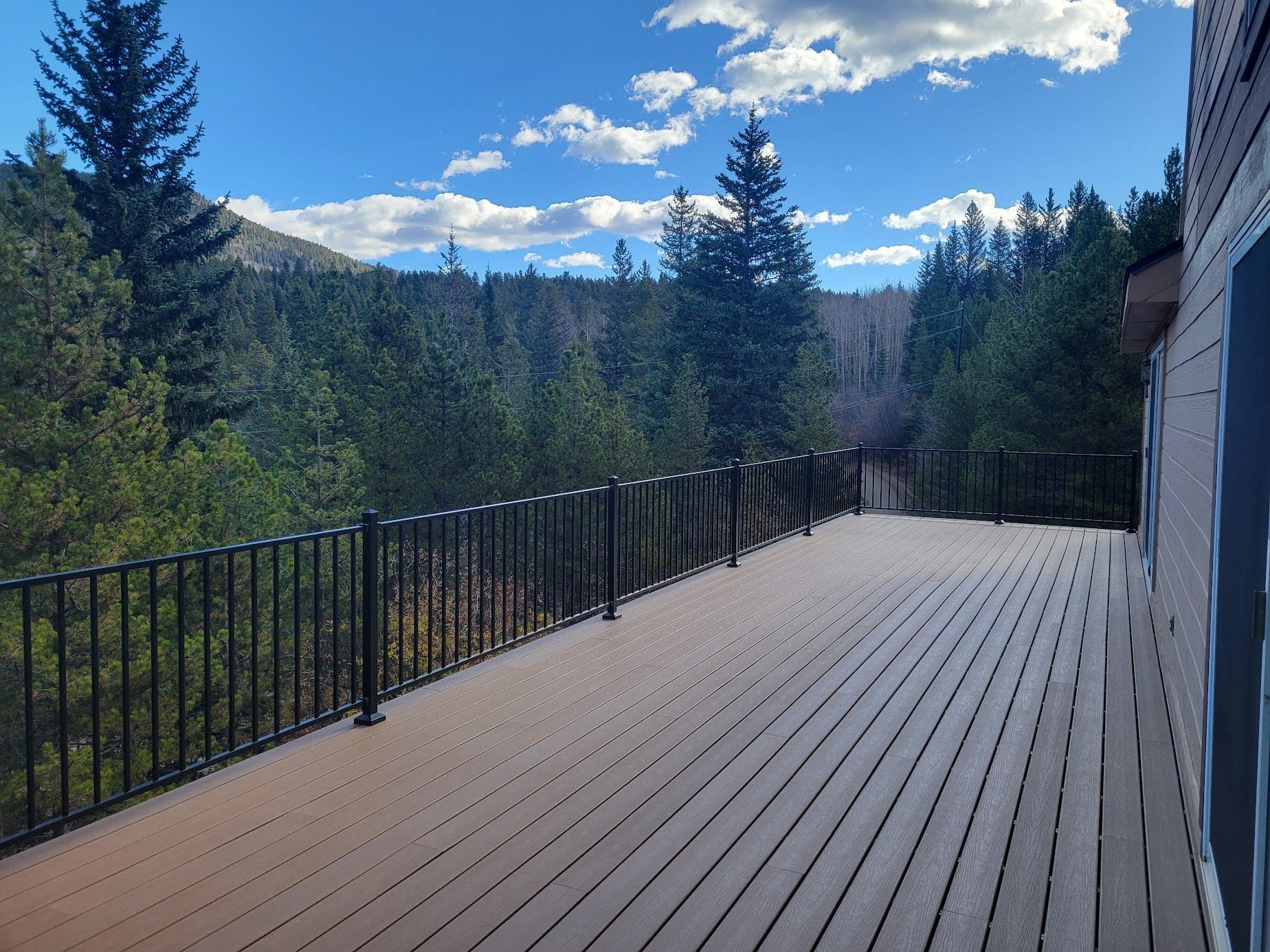 Wooden deck with black railing overlooking evergreen trees and mountains under a bright blue sky
