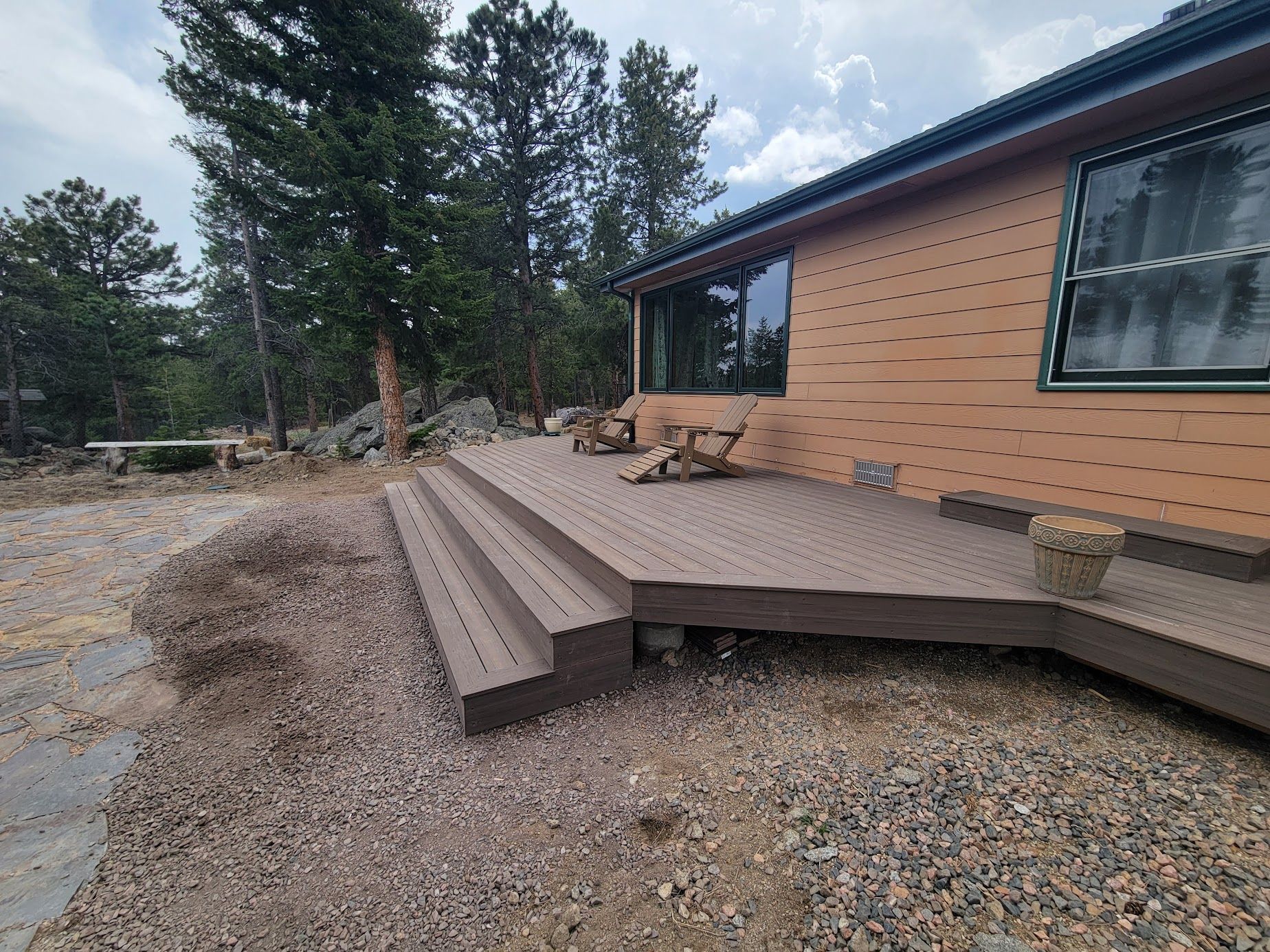 Wooden deck steps beside a tan house on a gravel lot, with trees in the background.