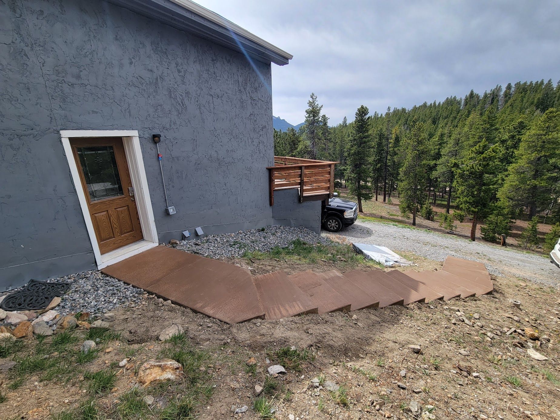 Concrete ramp beside a gray house leading to a side door, with trees and a gravel road in the background