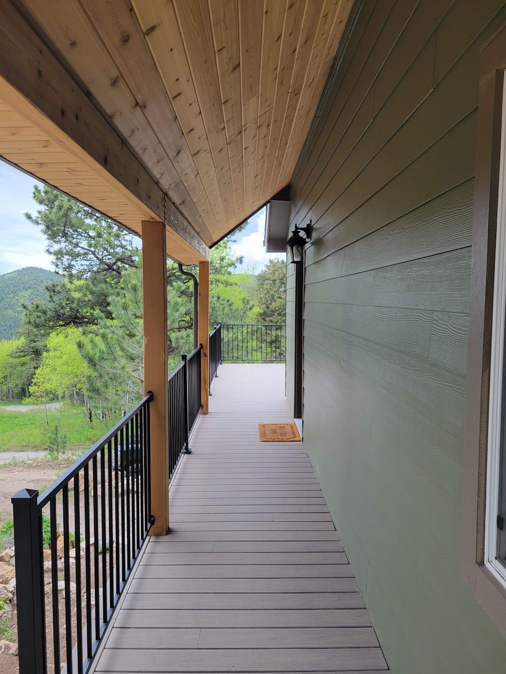 Covered porch with green siding, wood ceiling, and black railing overlooking a wooded hillside.