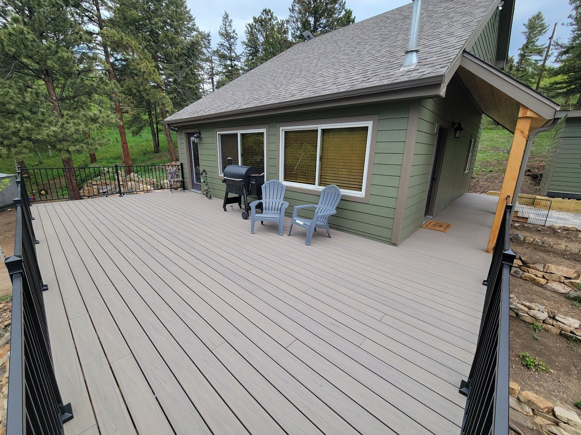 Wooden deck beside a green cabin with sliding windows, outdoor chairs, and a grill in a wooded yard