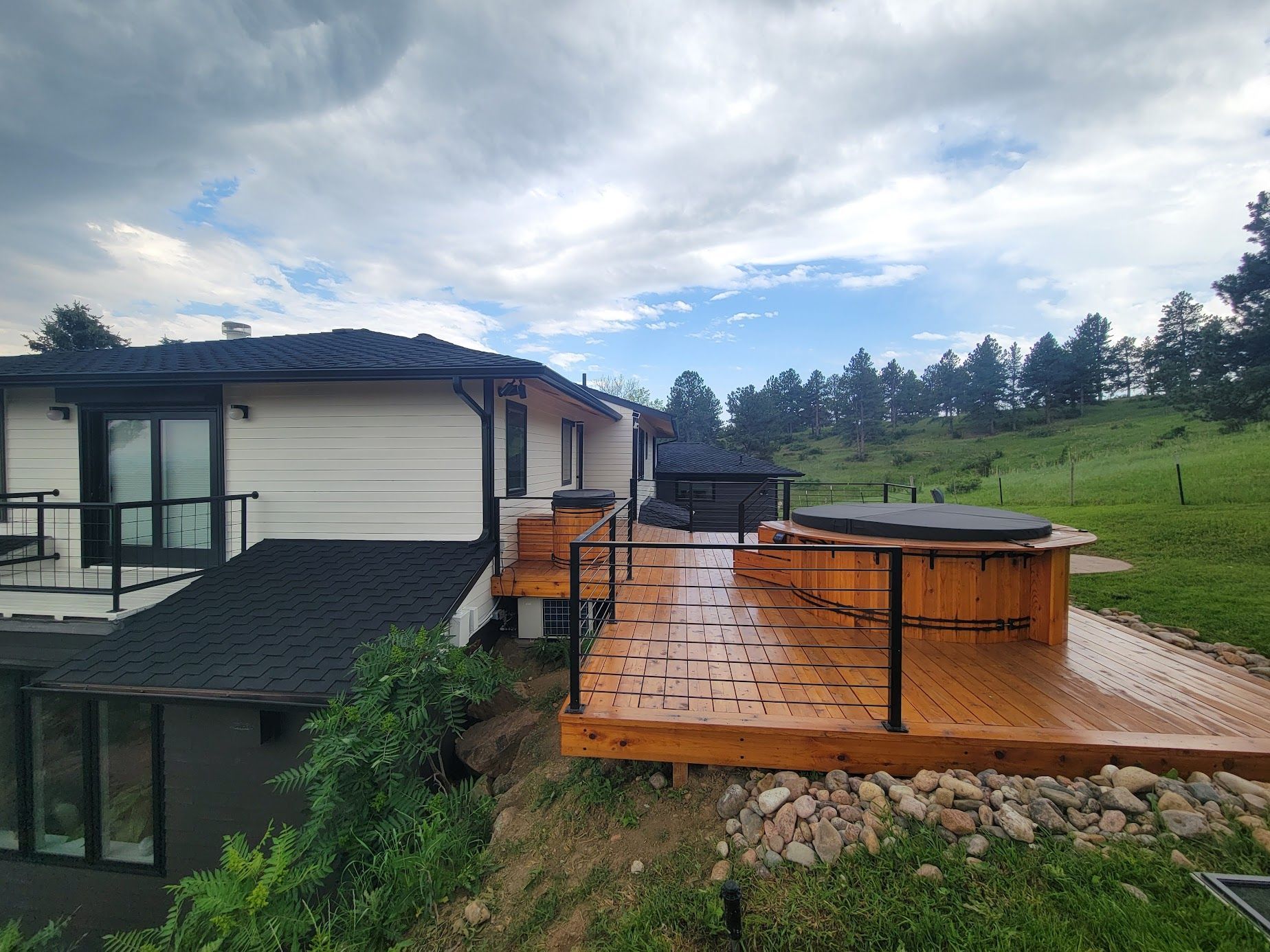 Modern house with wet wooden deck and hot tub on a grassy hillside under cloudy skies