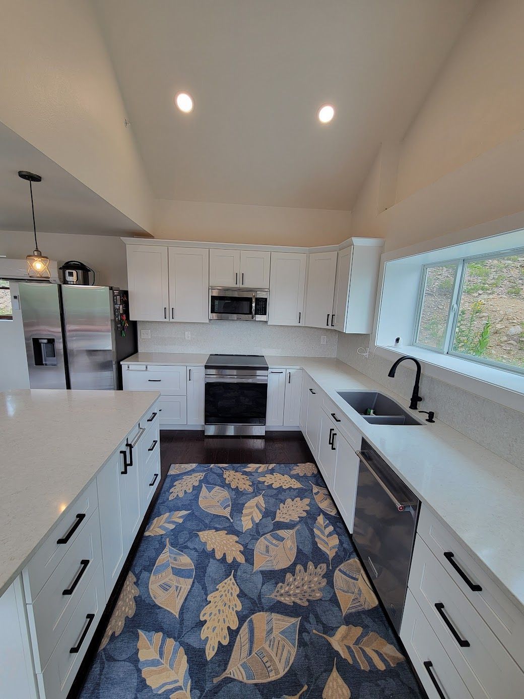 Bright kitchen with white cabinets, stainless steel appliances, patterned blue rug, and large window.