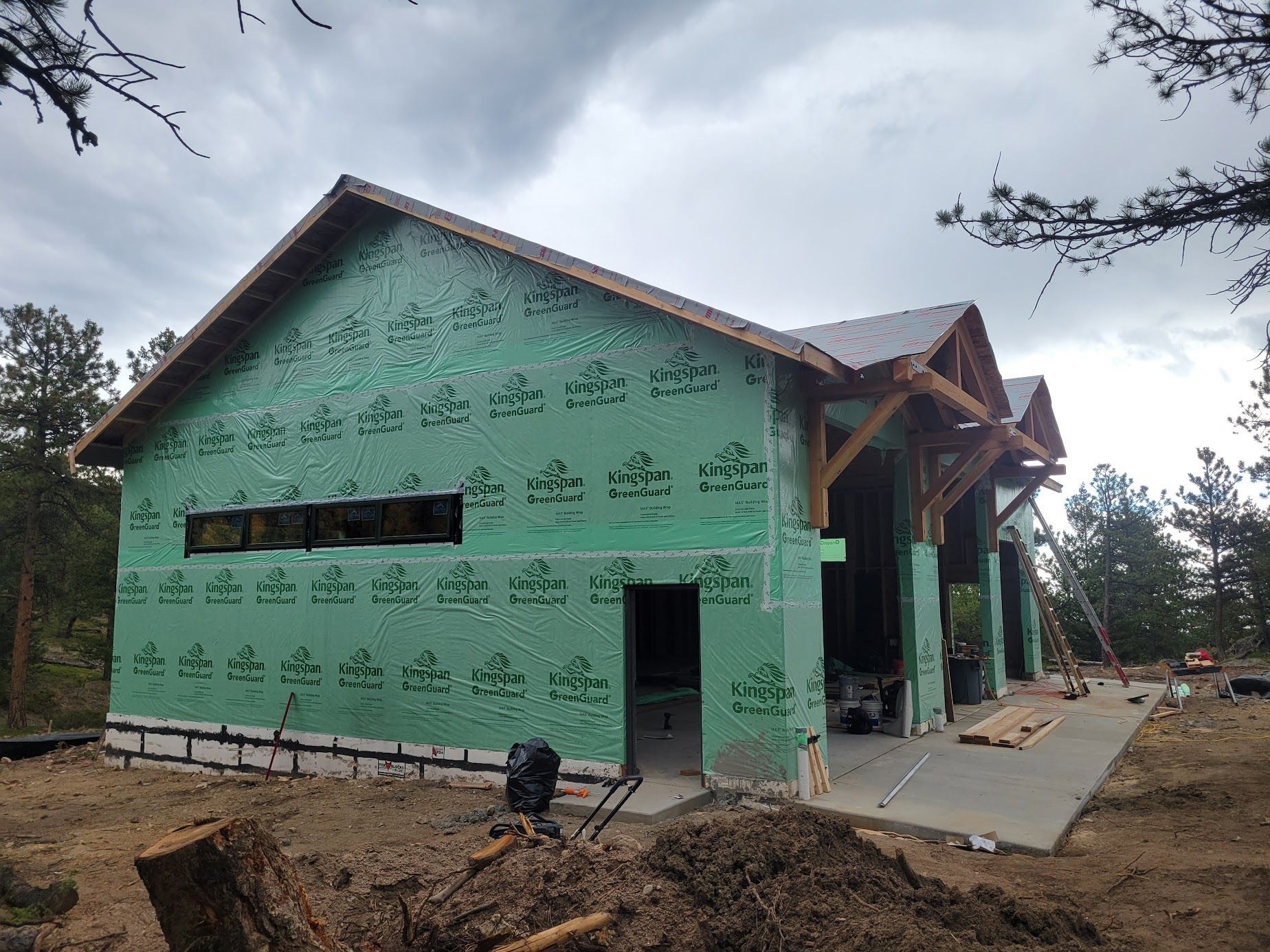 Green house under construction with exposed framing, a front porch, and bare dirt yard under cloudy skies