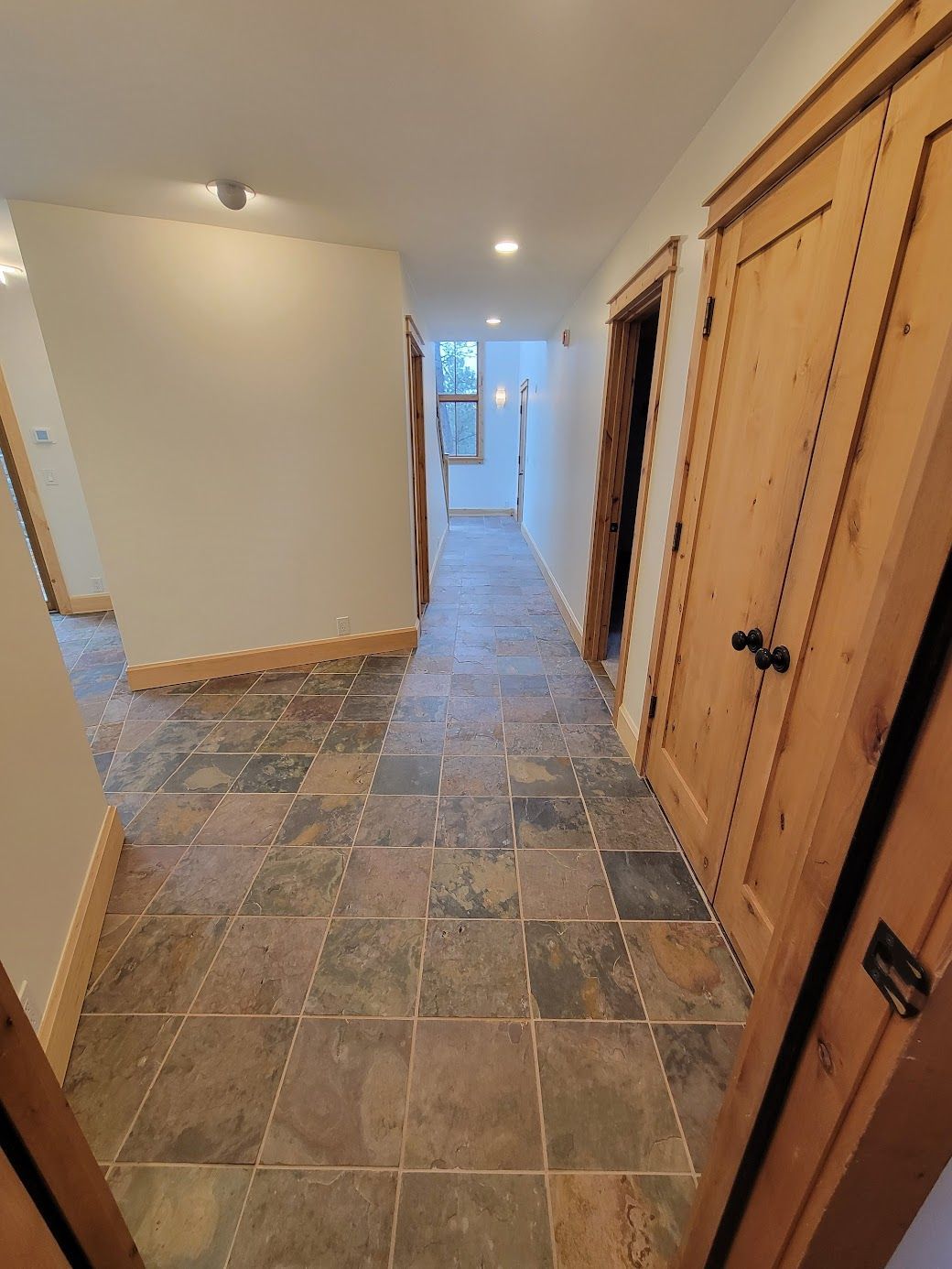 Narrow hallway with stone tile floor, wooden doors, and a bright doorway at the far end.