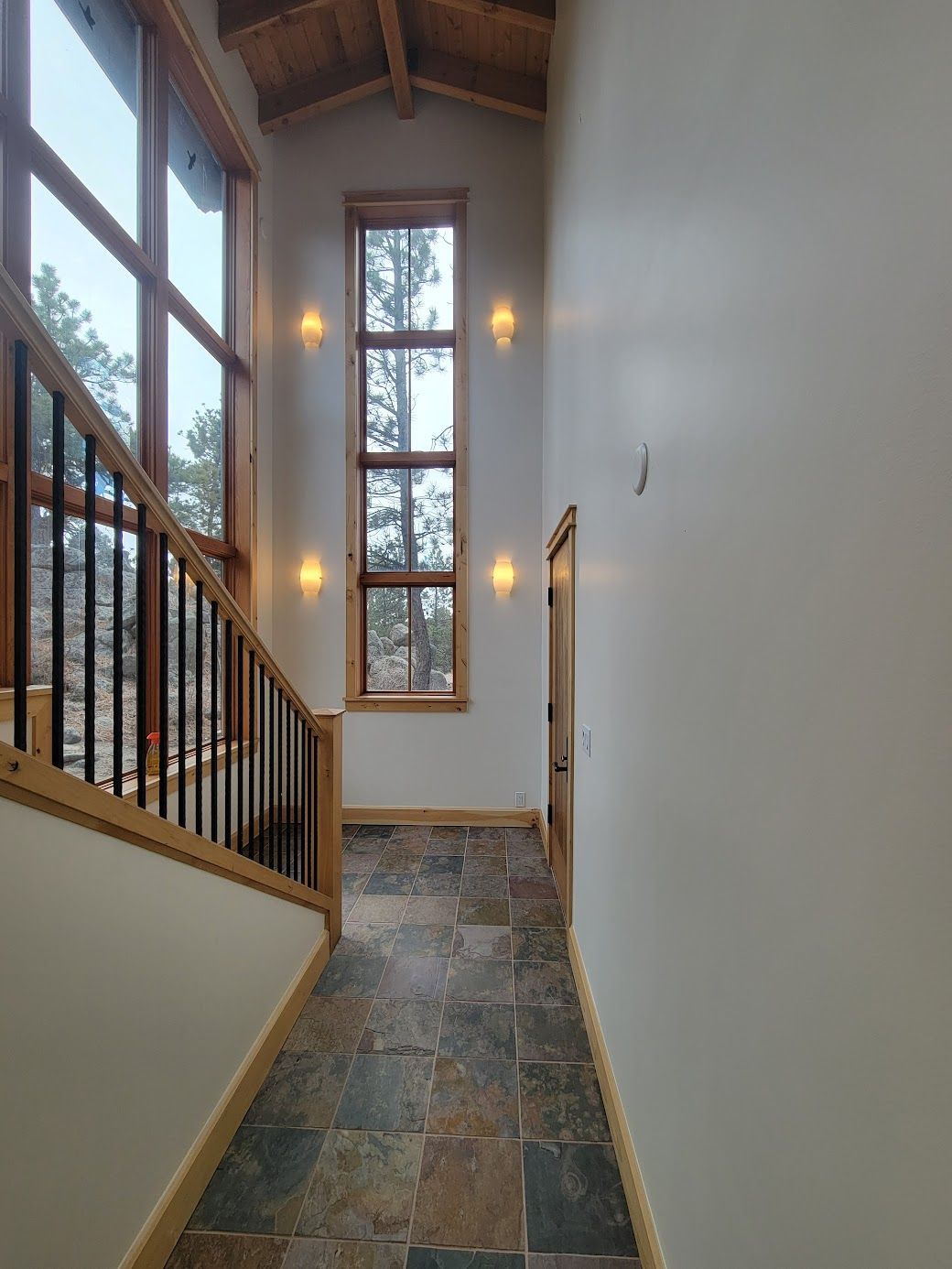Narrow stair landing with slate tile floor, tall window, white walls, wood railing, and warm wall sconces