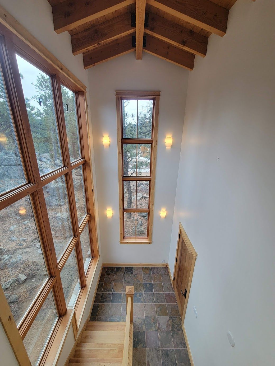 Narrow stairwell with tall windows, stone floor, and warm wall sconces beneath a wood-beamed ceiling
