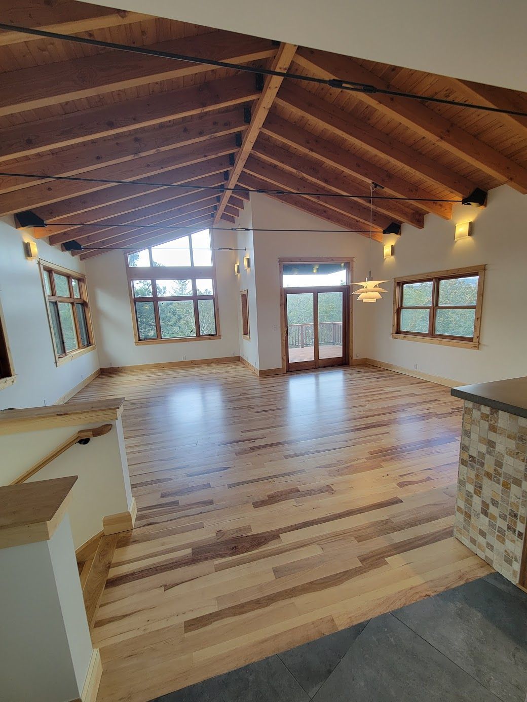 Empty living room with vaulted wood ceiling, hardwood floors, and large windows and glass doors.
