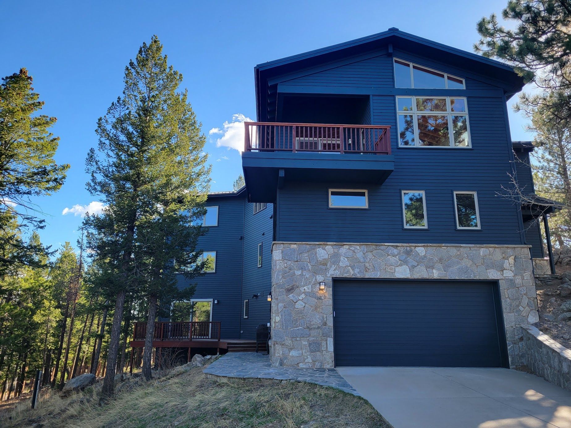 Modern dark-blue house with stone garage, red balcony, and pine trees under a clear sky