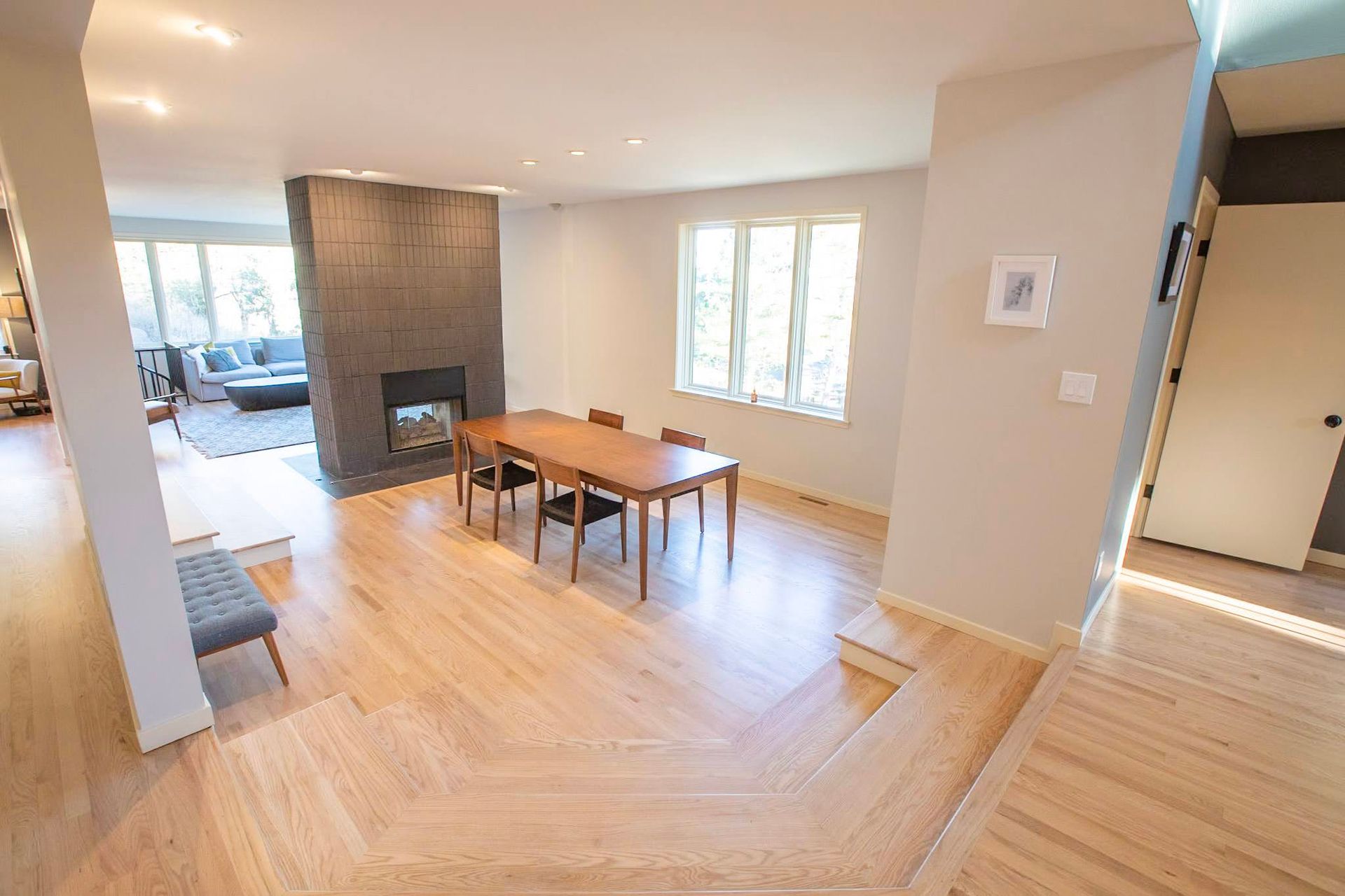 Bright dining room with wooden table, stone fireplace, and sunlit windows adjoining a living area