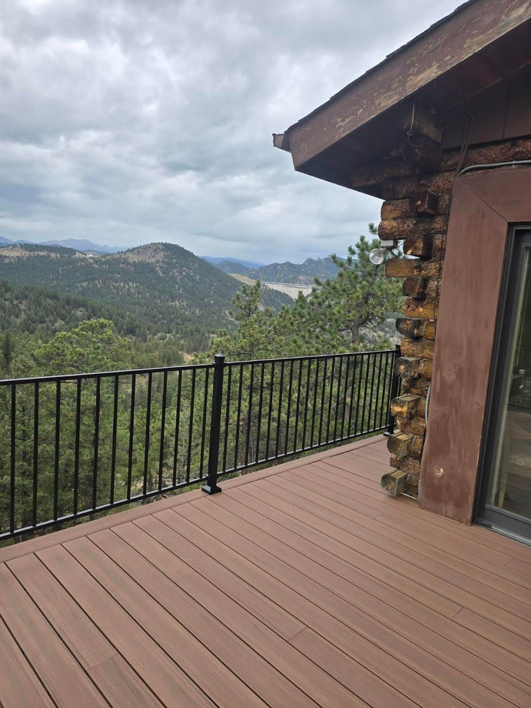 Wooden deck overlooking forested mountains under a cloudy sky, beside a rustic cabin.