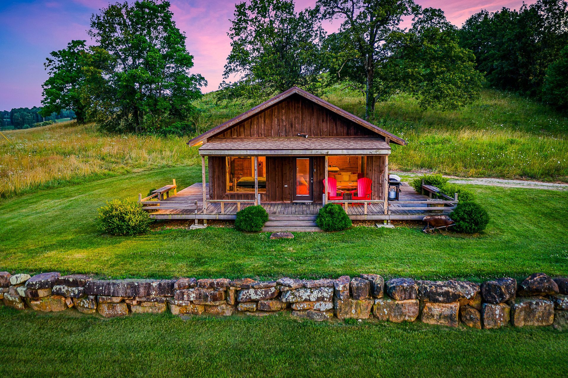 Small rustic cabin with a lit porch on a grassy hillside at dusk, surrounded by trees and stone edging