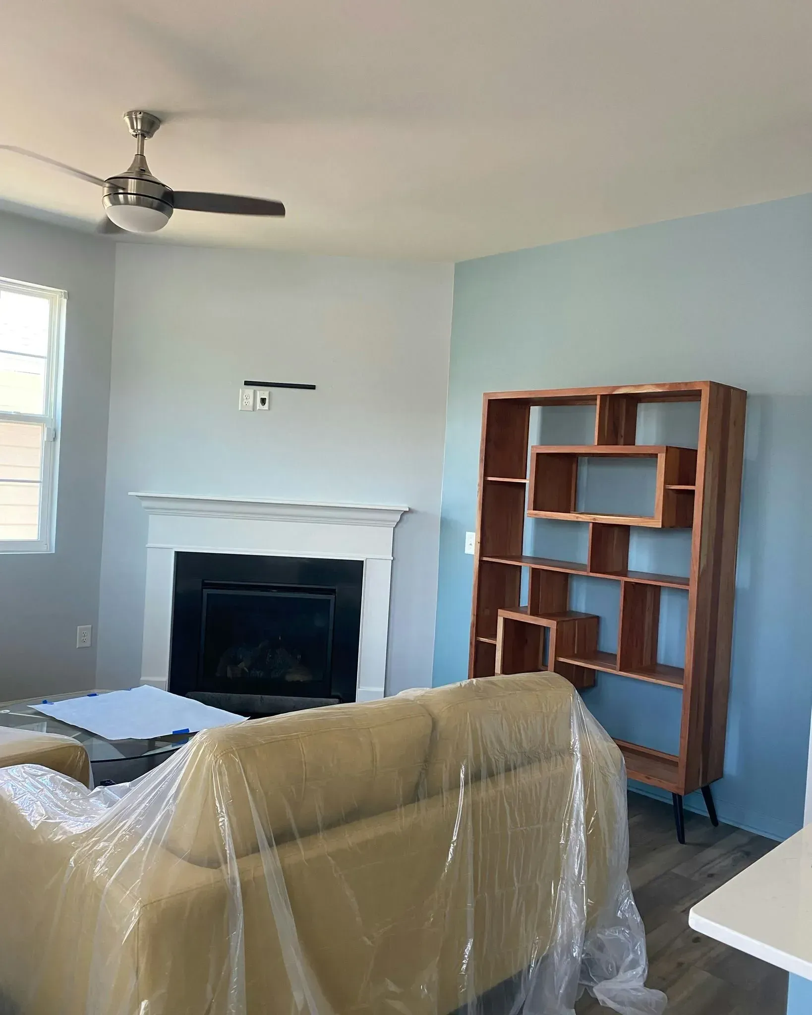 Living room with blue walls, white fireplace, ceiling fan, and wooden bookshelf; sofa wrapped in plastic in foreground