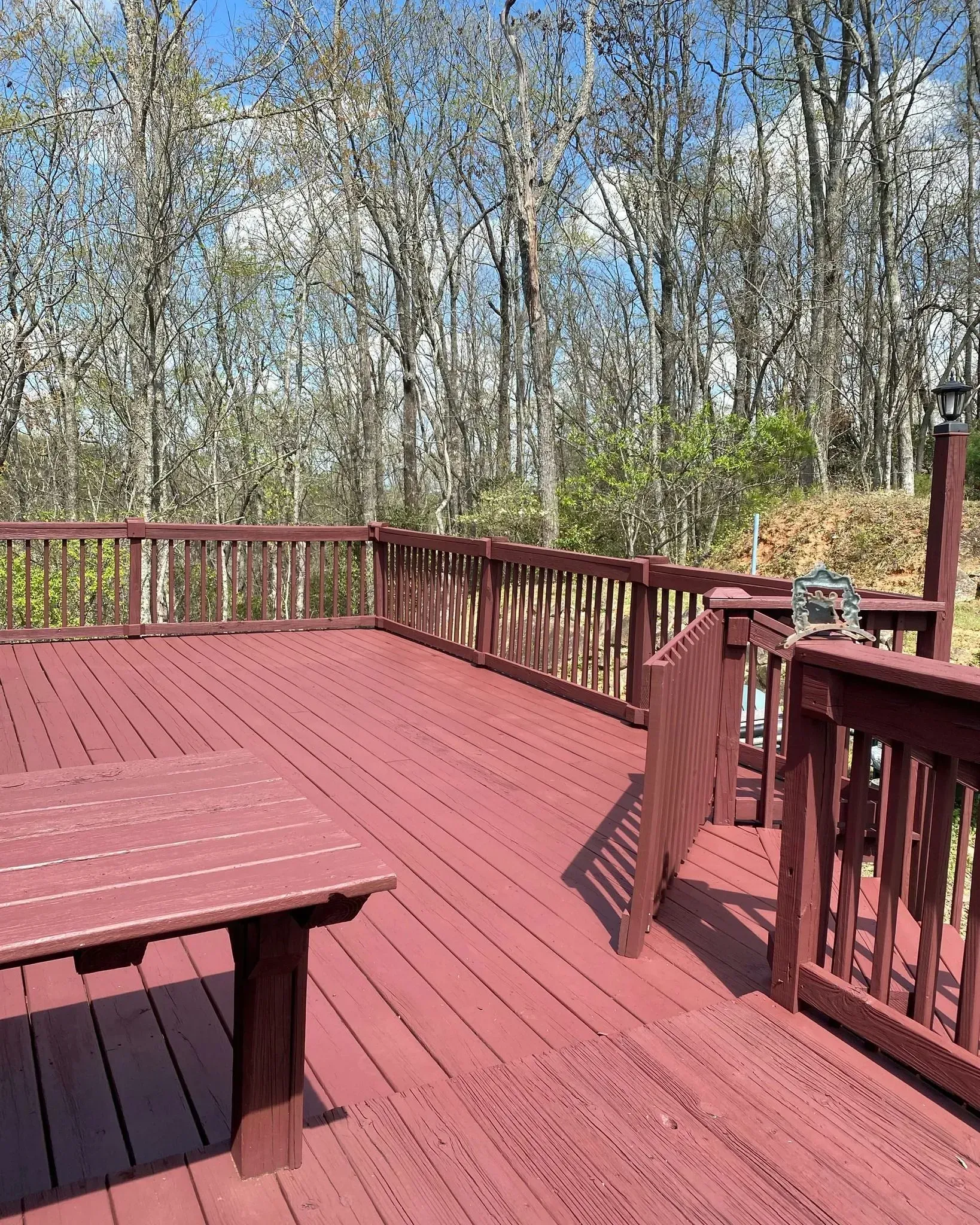 Red wooden deck with railing and bench overlooking leafless trees under a blue sky.