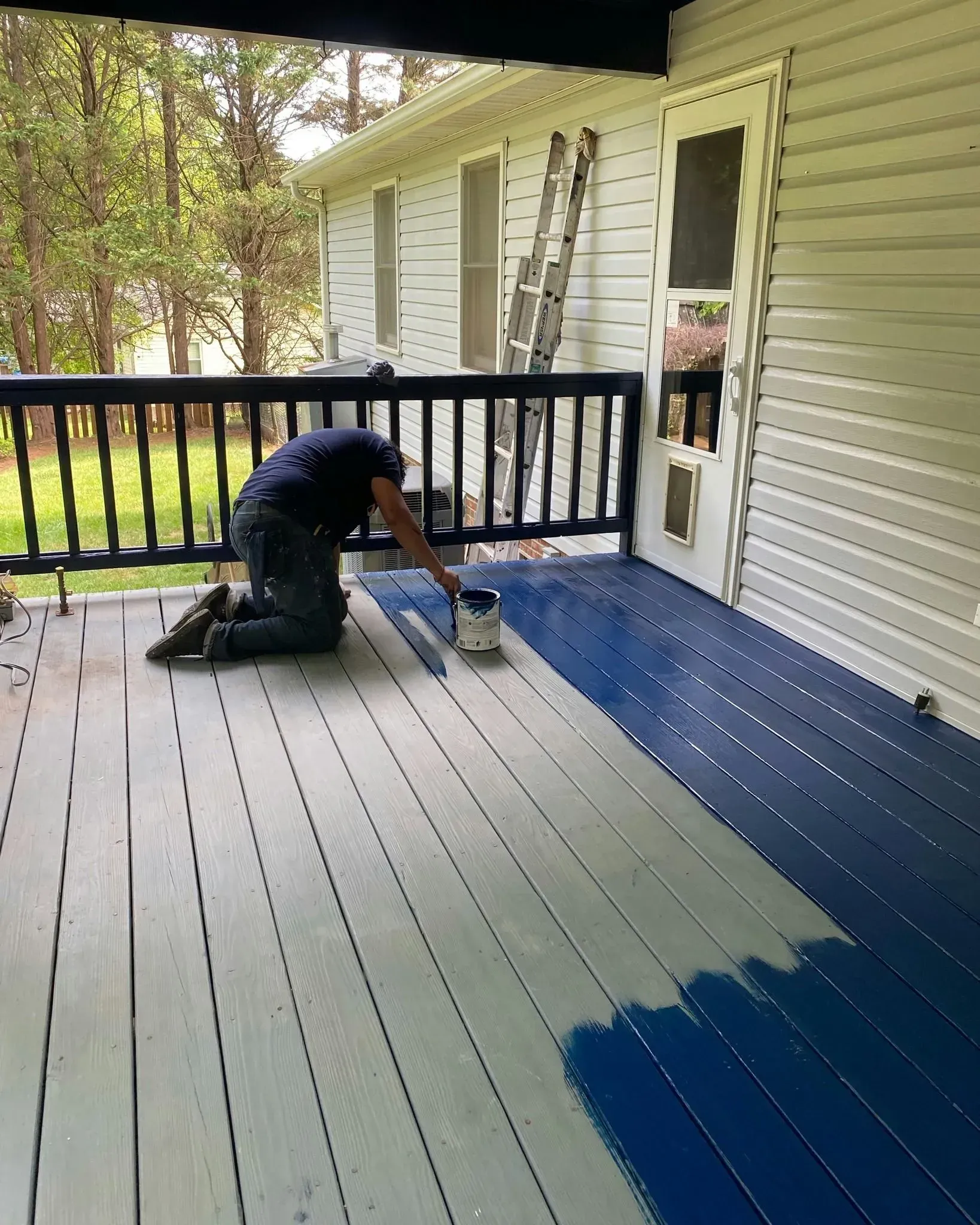 Person painting a porch floor blue beside a ladder and white siding