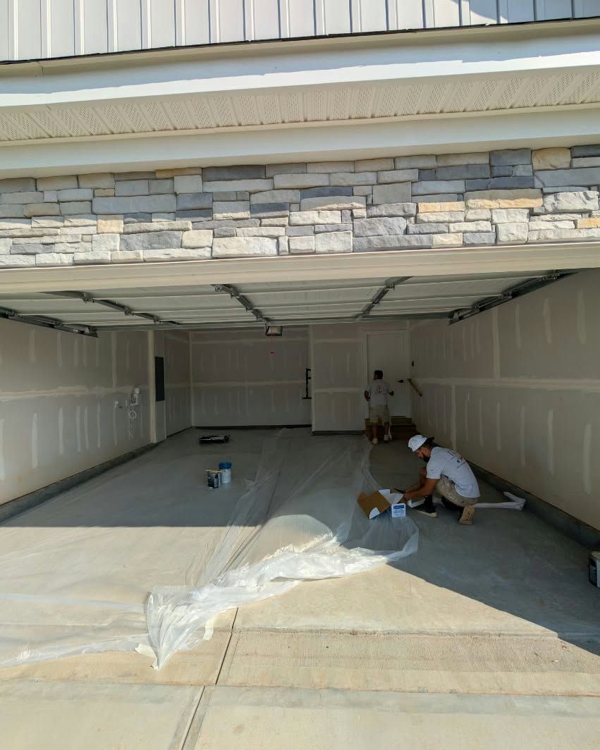 Two workers laying flooring inside an empty garage with drywall walls and an open door