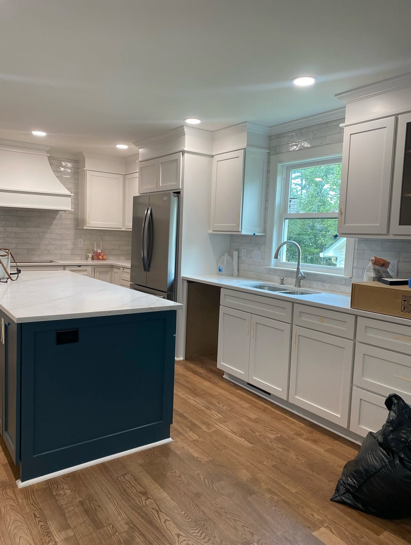 Bright kitchen with white cabinets, blue island, stainless fridge, wood floor, and a window over the sink.
