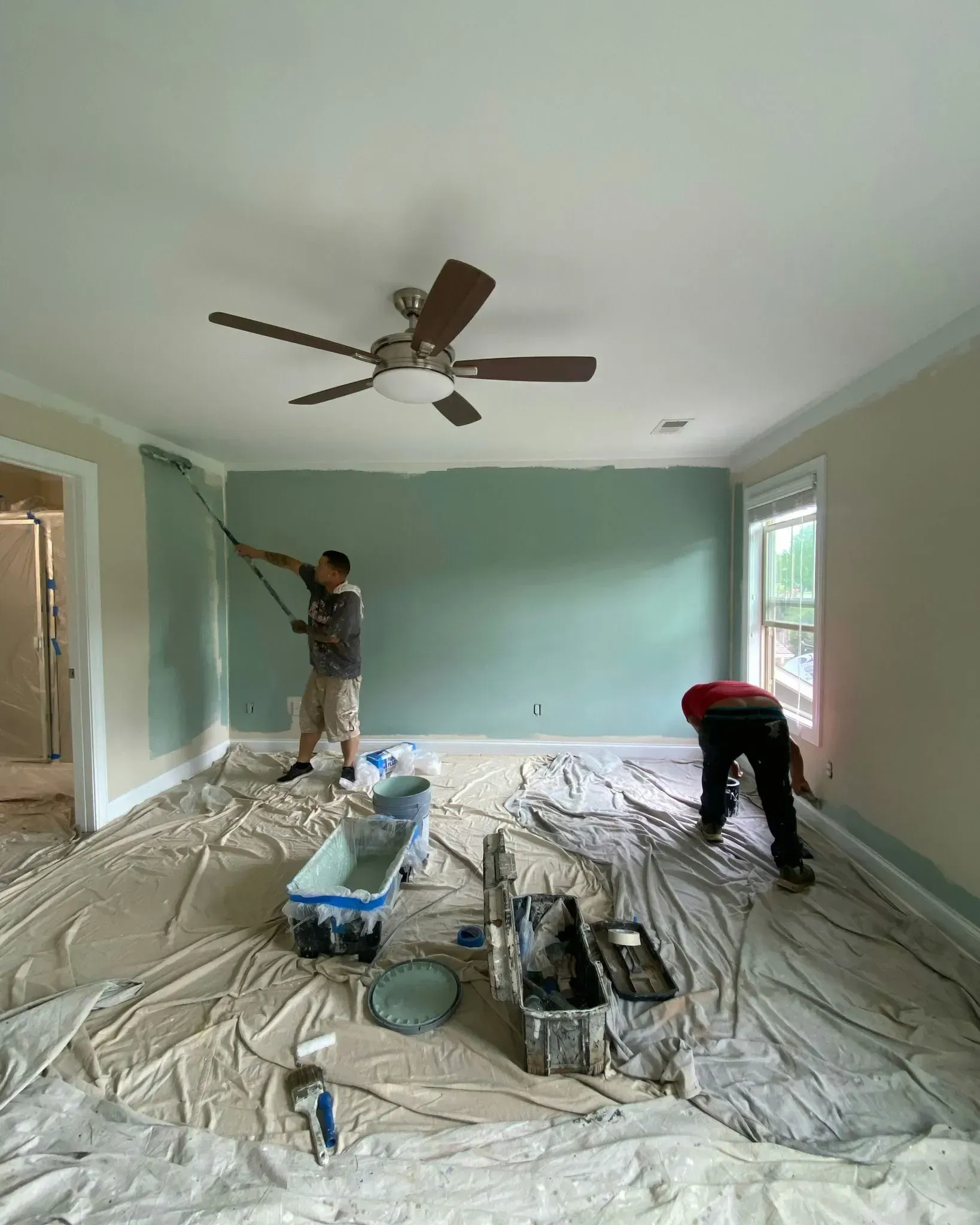 Two people painting a room with teal walls, covered floor, and a ceiling fan overhead.