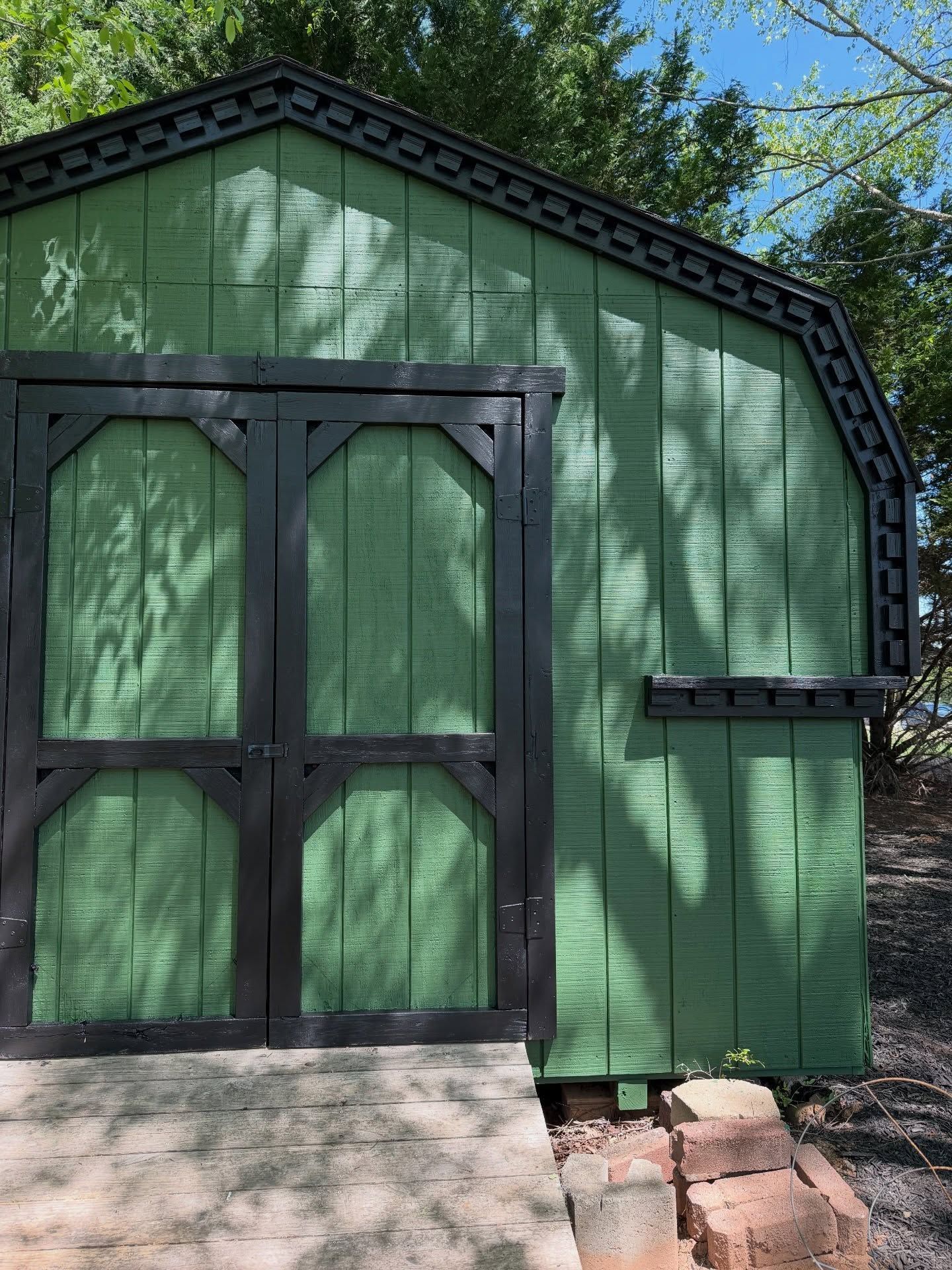 Green garden shed with double black doors and a small side shelf in dappled sunlight