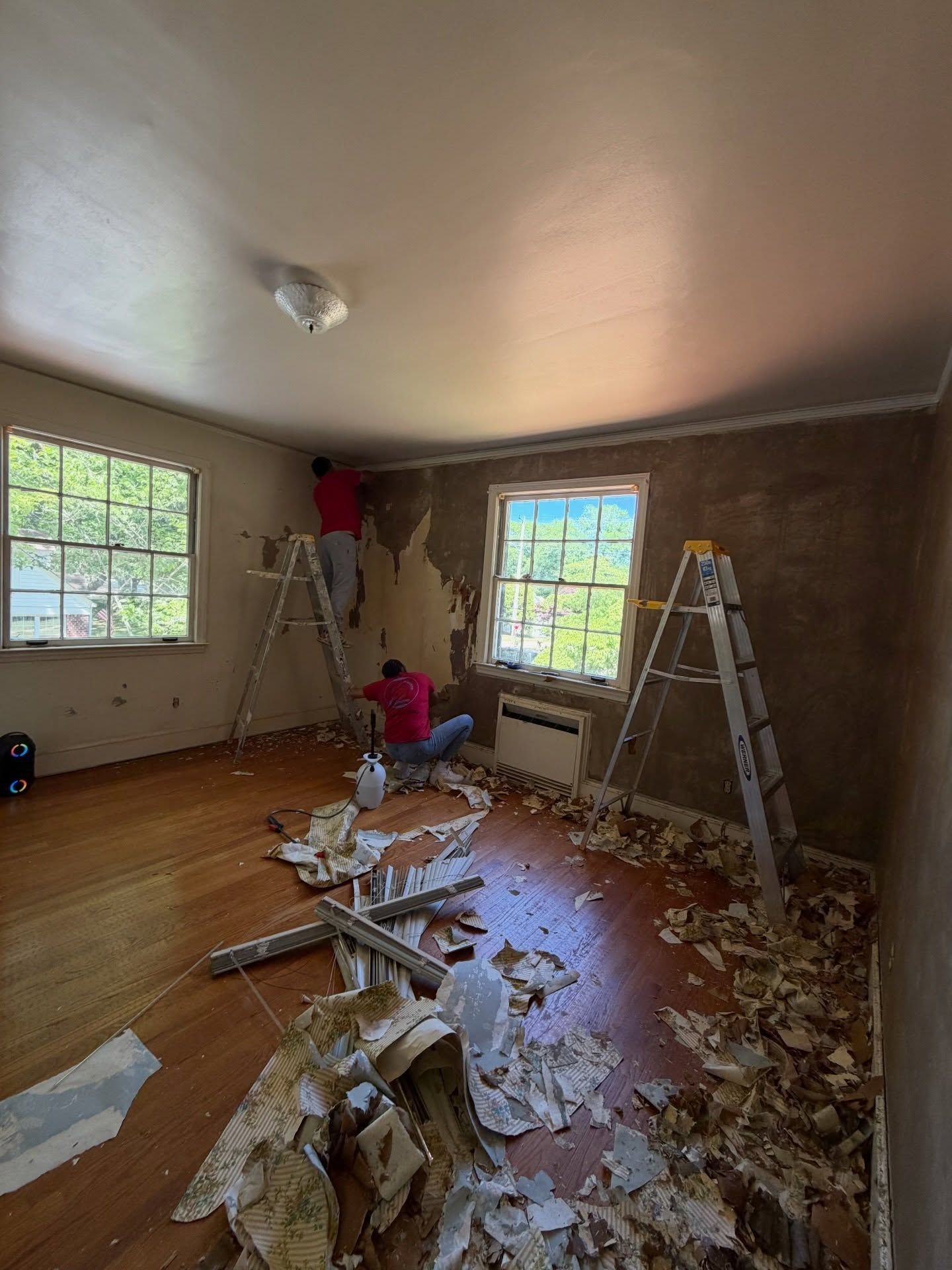 Renovation-in-progress room with torn wallpaper, debris on floor, and a ladder by two windows.