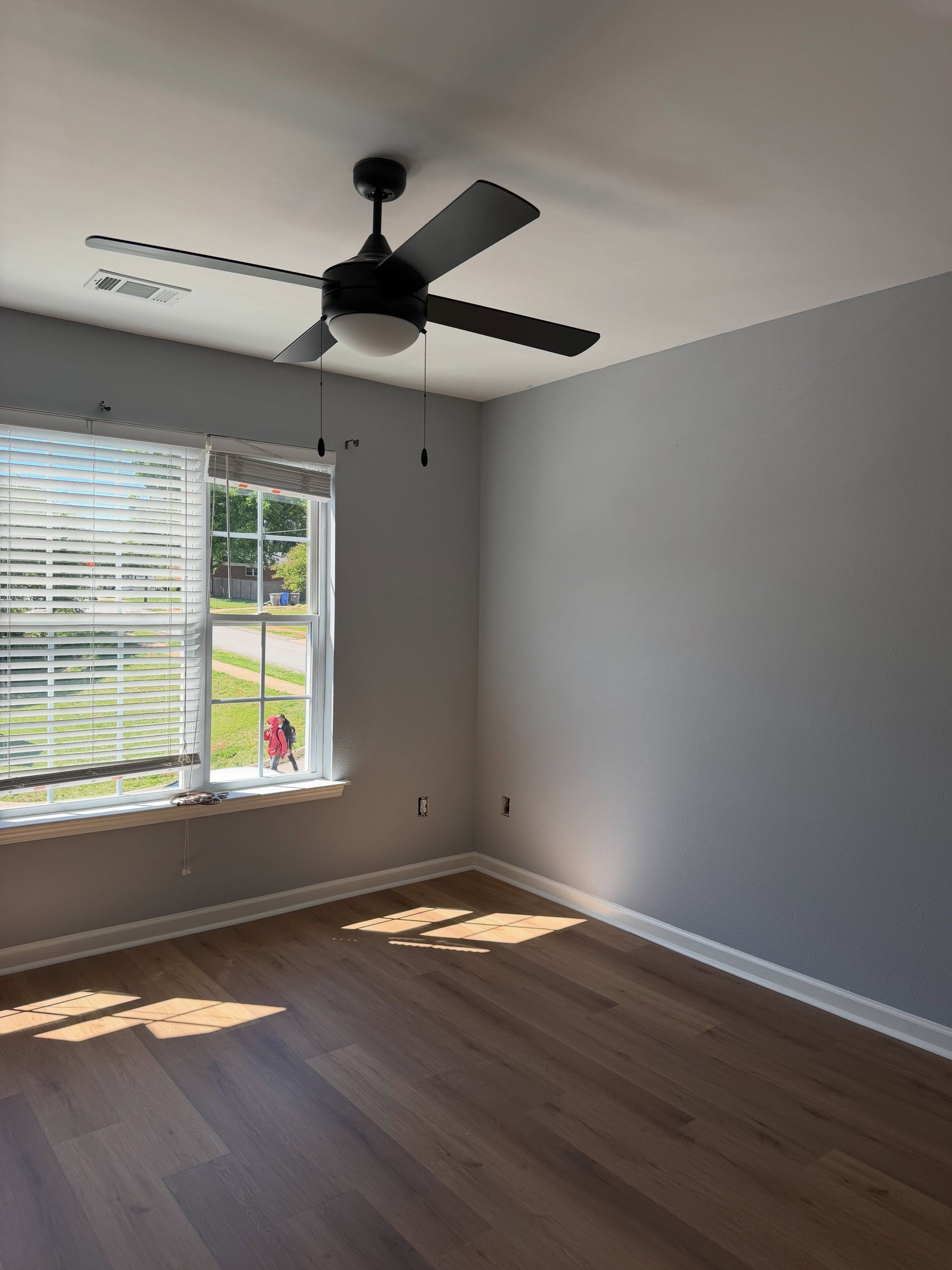 Empty room with gray walls, wood floor, ceiling fan, and sunlight through a window with blinds.