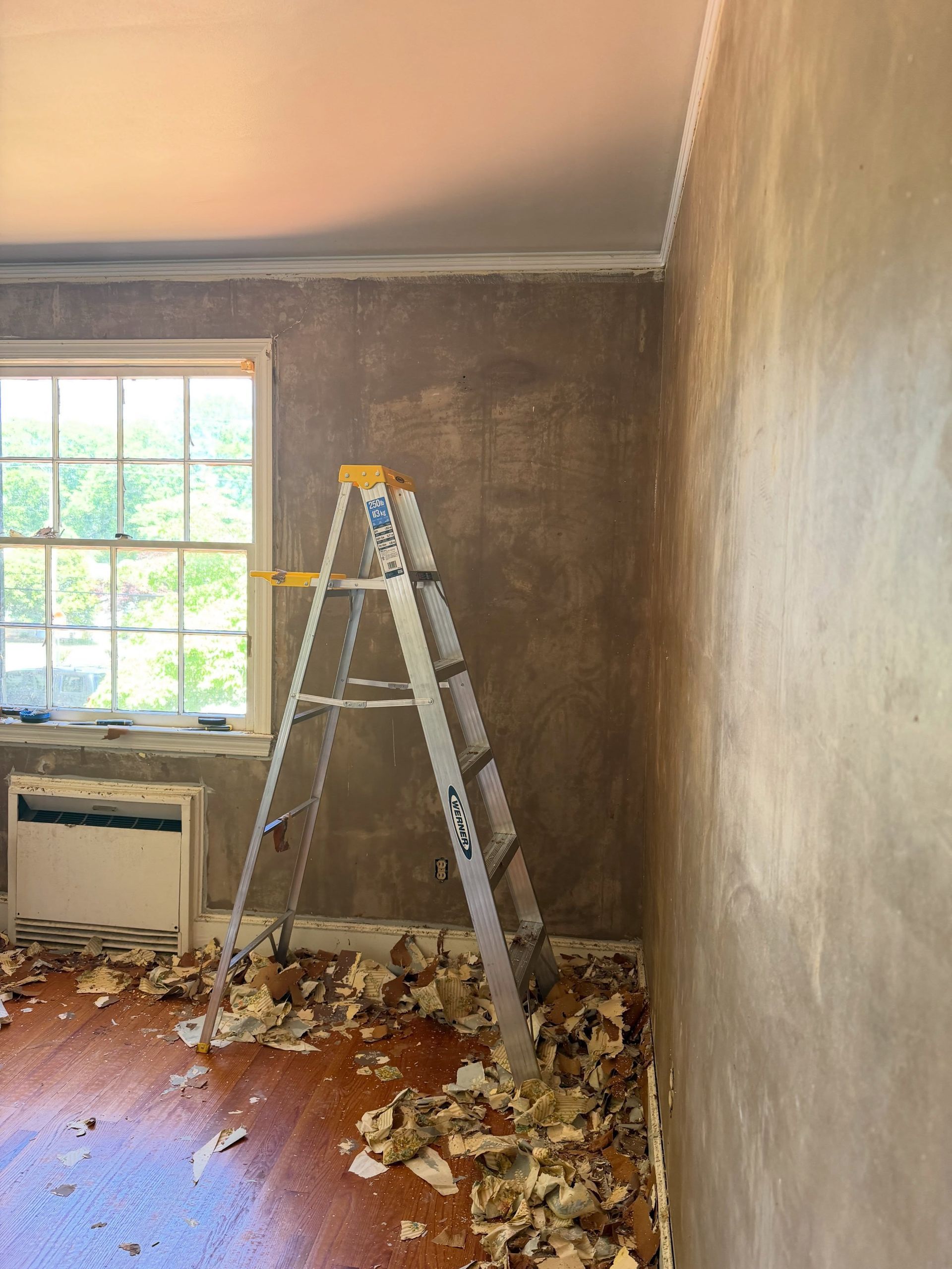 Room under renovation with ladder, debris on floor, gray walls, and window with AC unit