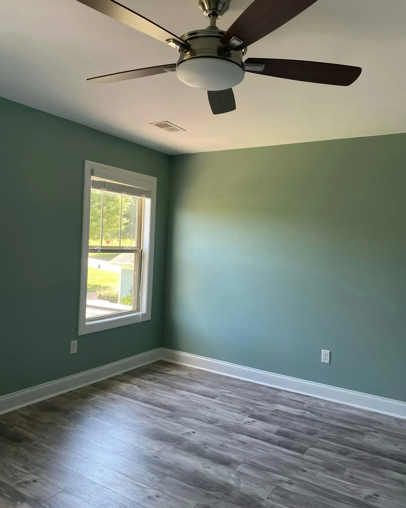 Empty room with sage green walls, gray wood flooring, a window, and a ceiling fan.