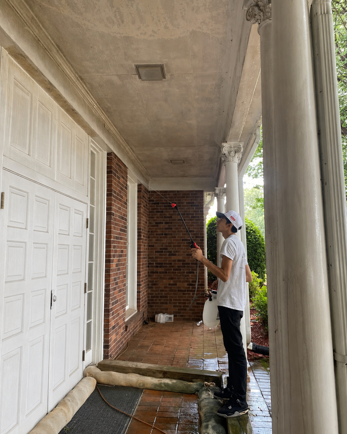 Person painting a porch ceiling beside white garage doors and brick columns with a roller pole