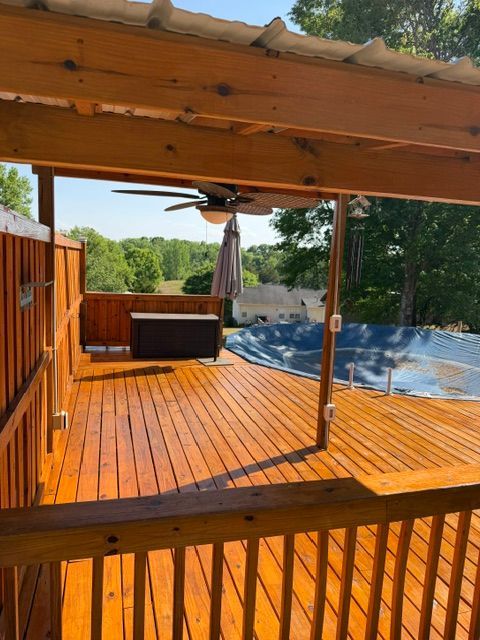 Covered wooden deck with railing, ceiling fan, patio furniture, and a closed umbrella overlooking trees and a pool area