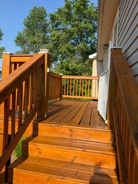 Wooden deck stairs and railing leading to a sunlit porch beside a white house, with trees beyond.