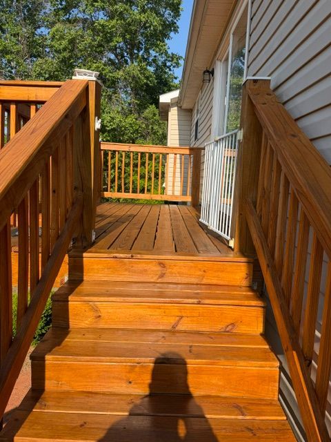 Wooden deck stairs and railings beside a house, leading to a sunlit porch with trees beyond.