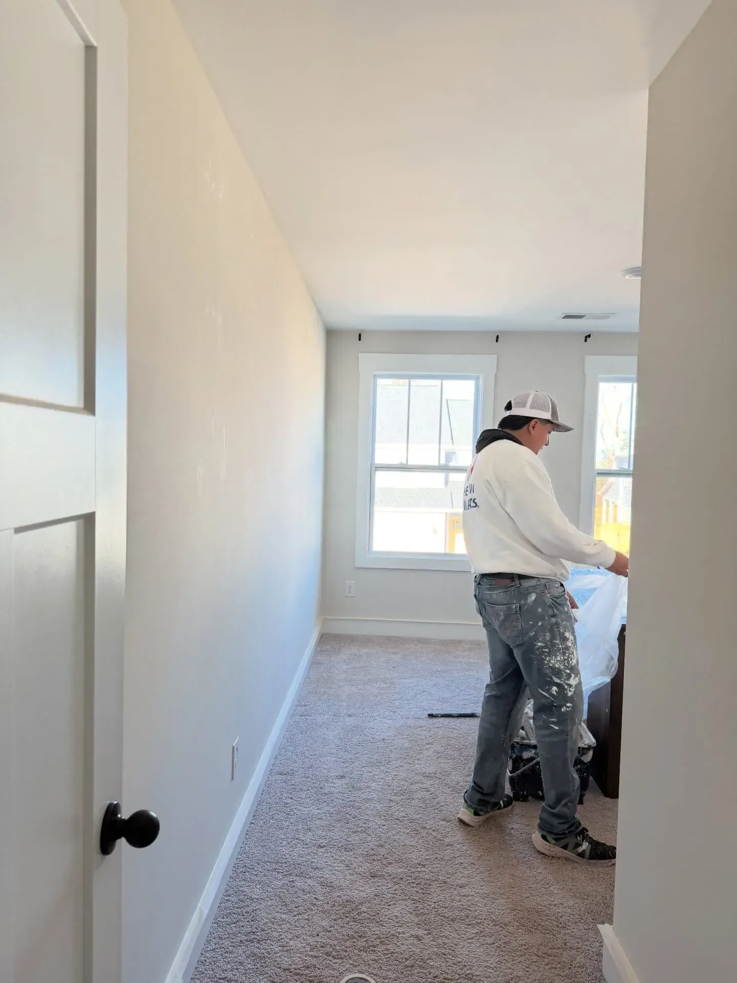 Worker painting a narrow bedroom with beige carpet and white walls, near two windows.