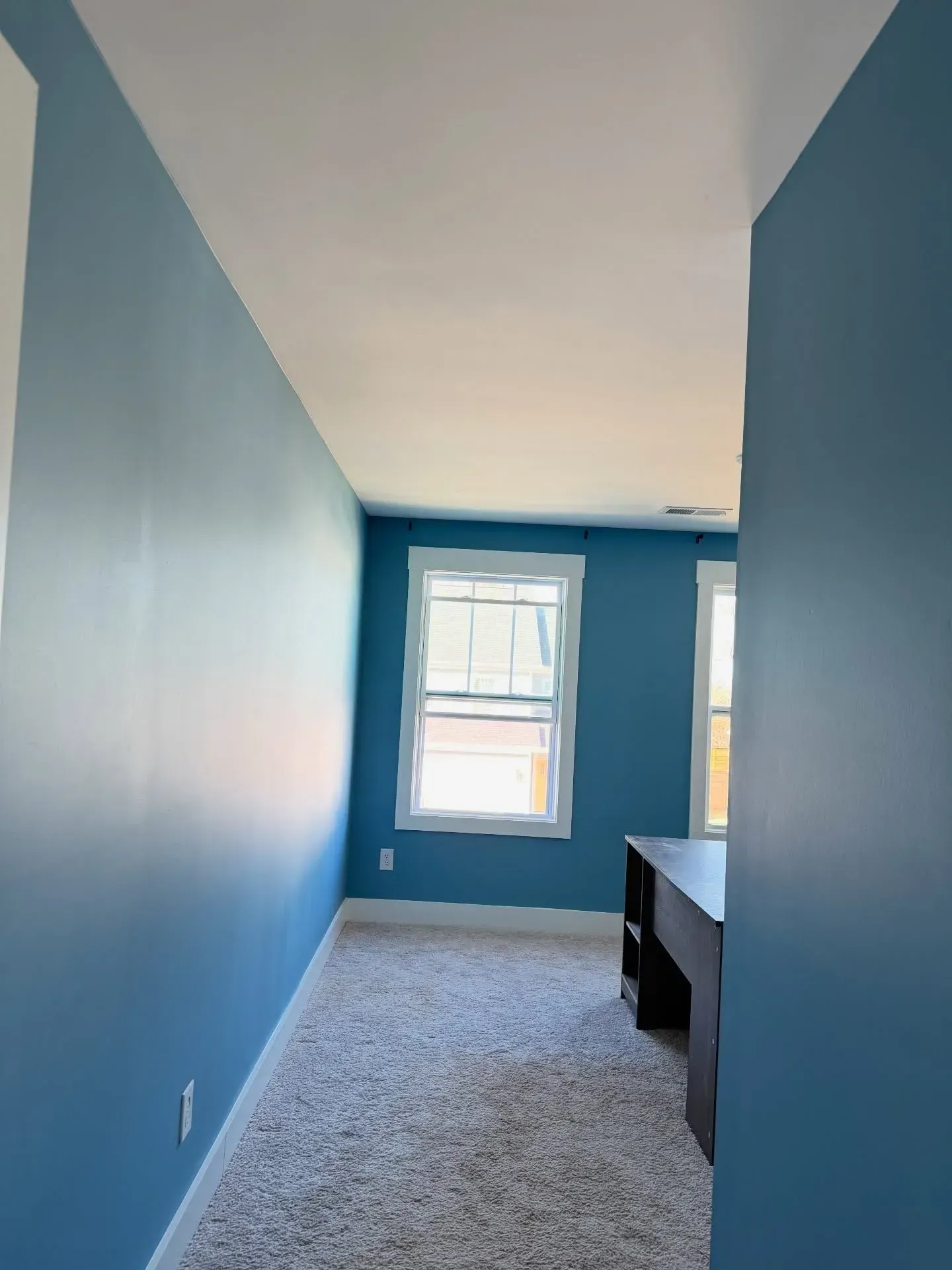 Empty blue room with a window, white ceiling, carpet, and a dark desk against the wall