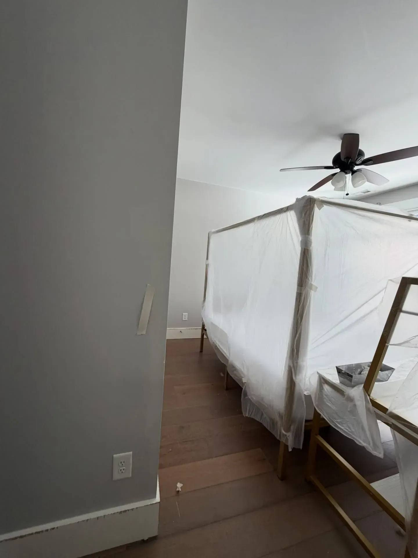 Bedroom with a white canopy bed under plastic wrap, wood floor, gray wall, and ceiling fan.