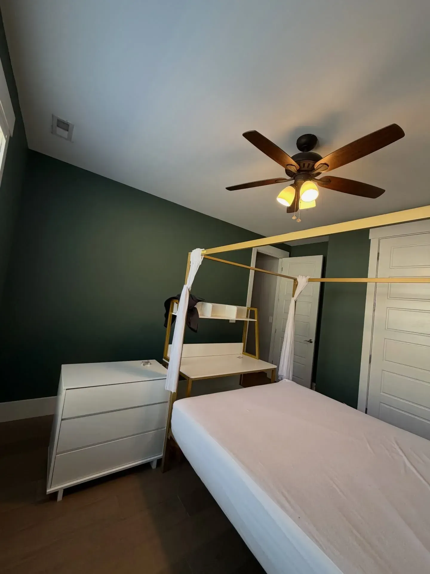 Bright bedroom with a four-poster bed, white dresser, green walls, and a ceiling fan with lights.